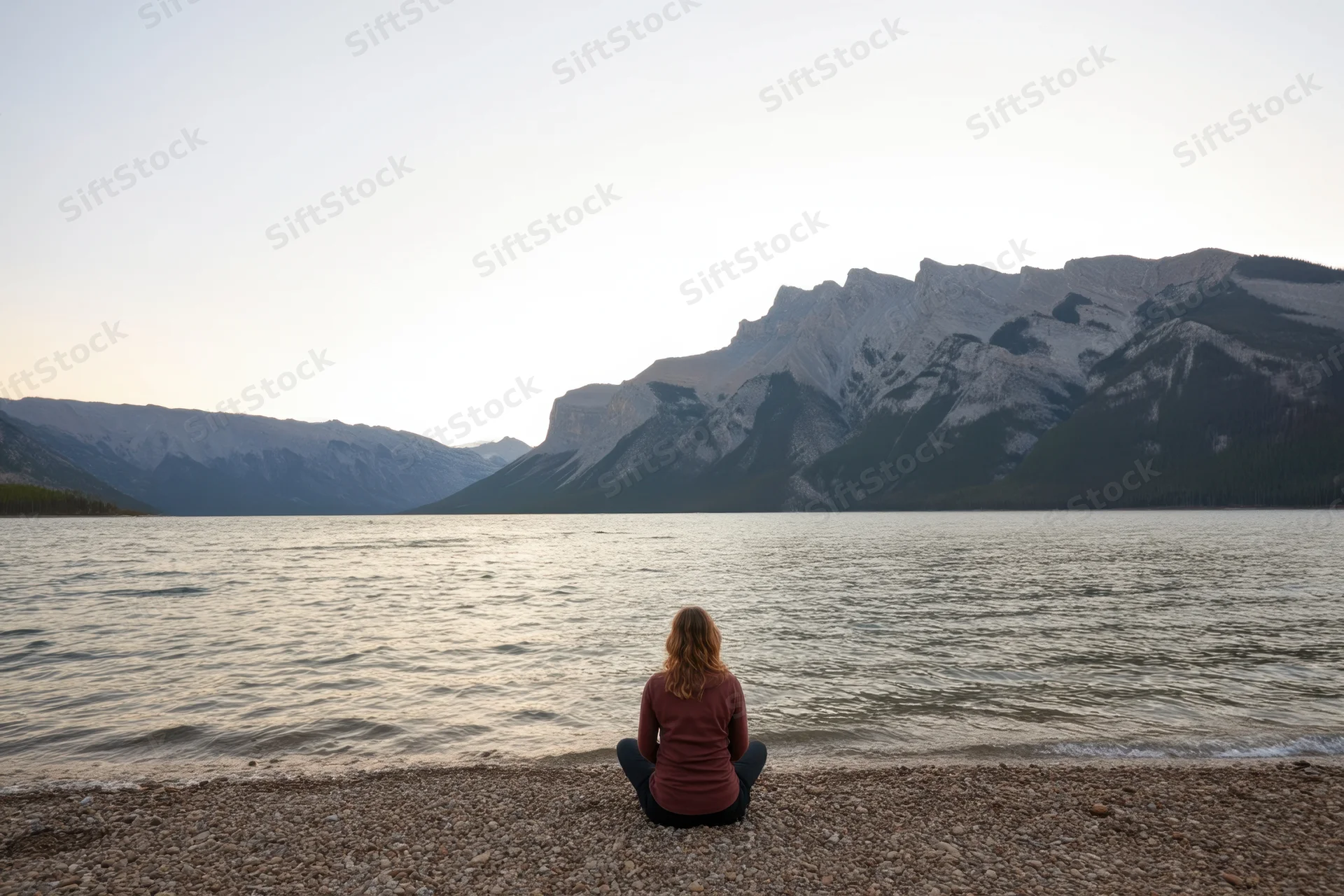 Woman sitting cross-legged on pebbled shore gazing at lake and mountains at sunset