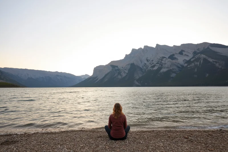 Woman sitting cross-legged on pebbled shore gazing at lake and mountains at sunset