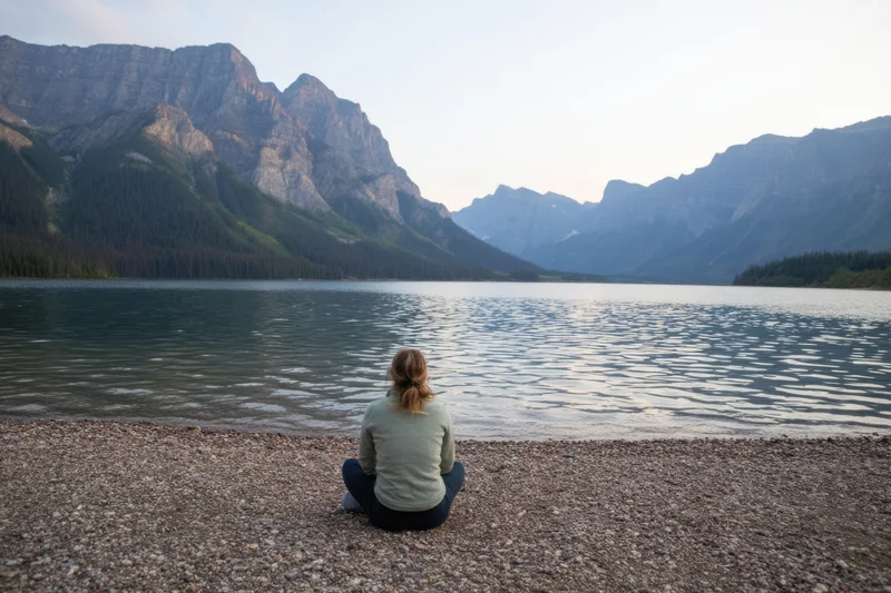 Woman sitting cross-legged on pebbled shore gazing at lake and mountains at twilight