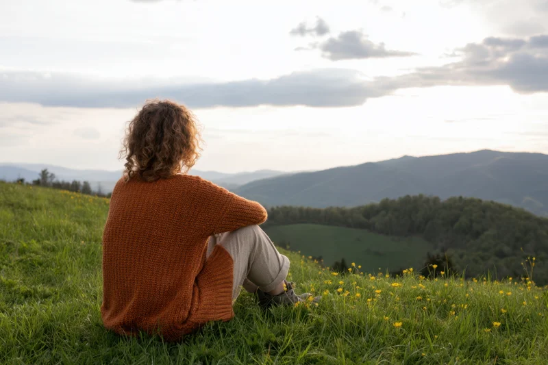 Woman sitting on grassy hillside with yellow flowers overlooking mountains at sunset