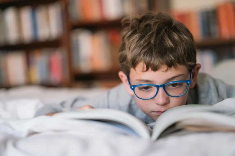 Young boy with blue-framed glasses reading an open book while lying on a bed in a bedroom with bookshelves behind him