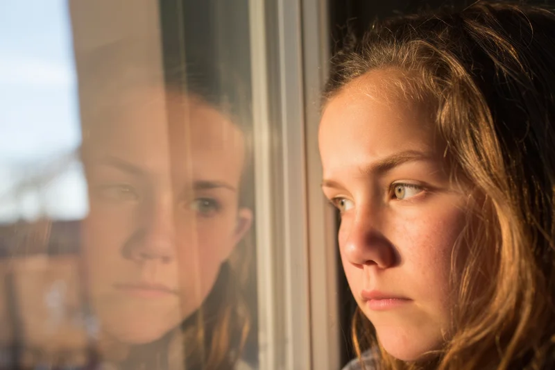 Young girl gazing out of window with her reflection visible on glass