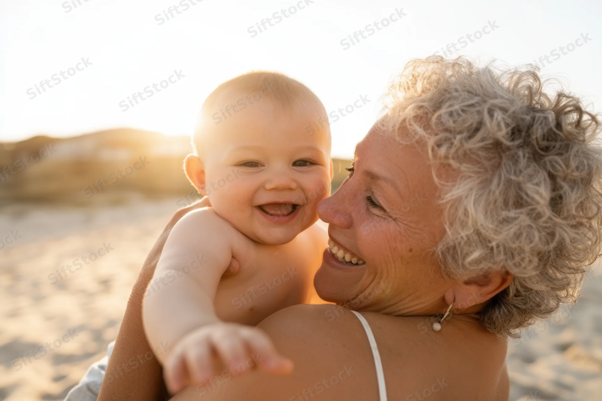 Elderly woman holding laughing baby on beach at sunset