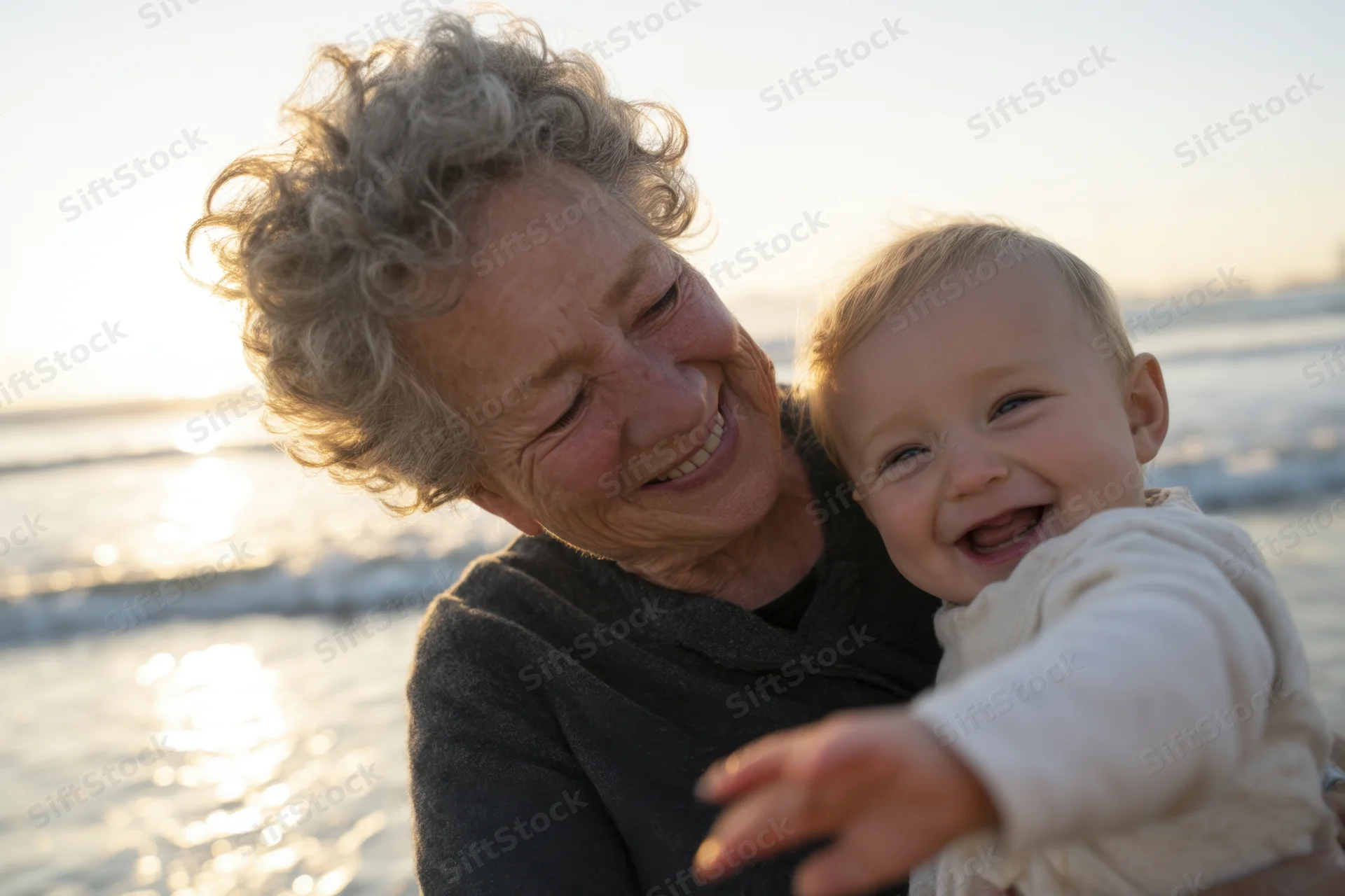 Elderly woman holding laughing baby on beach at sunset