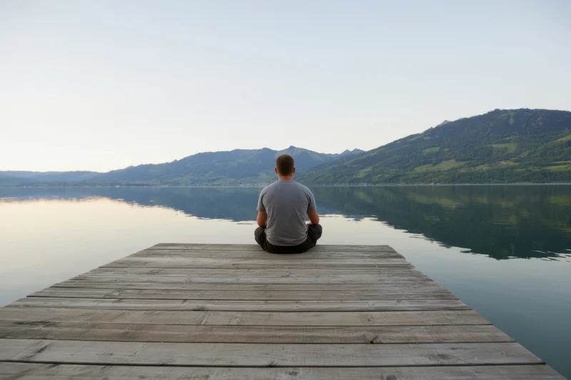 Man sitting cross-legged on wooden dock facing calm lake with mountains in distance