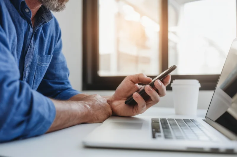 Man using smartphone while sitting at desk with laptop in casual office setting