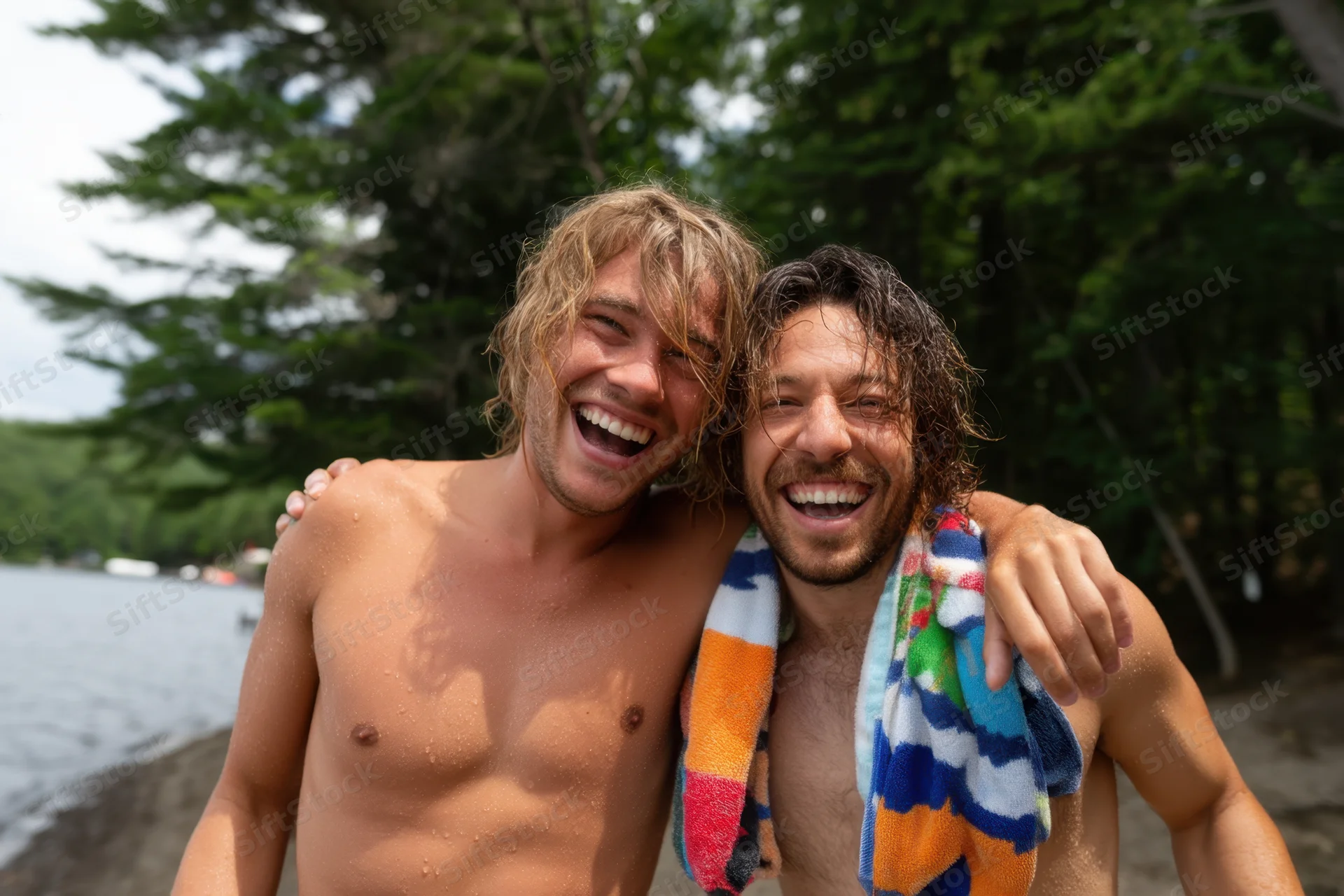 Two shirtless men laughing together on a lakeshore with trees behind them