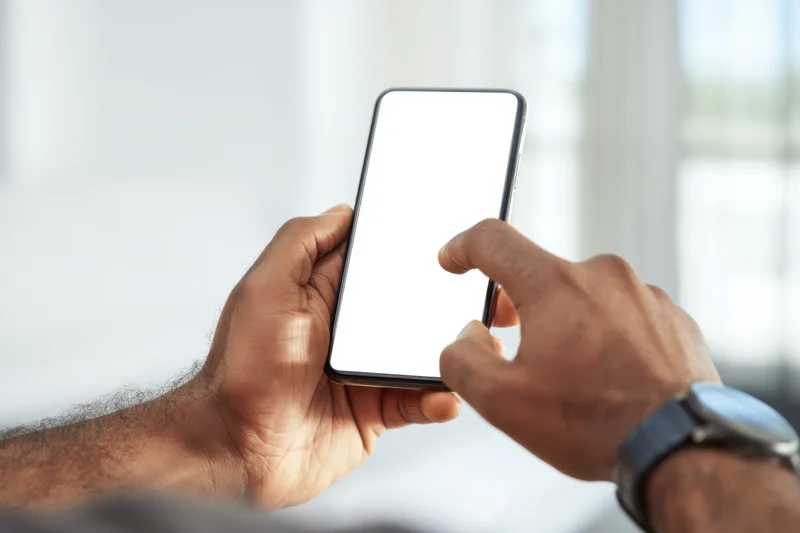 Hands holding smartphone with blank white screen, one finger touching display, indoors near window
