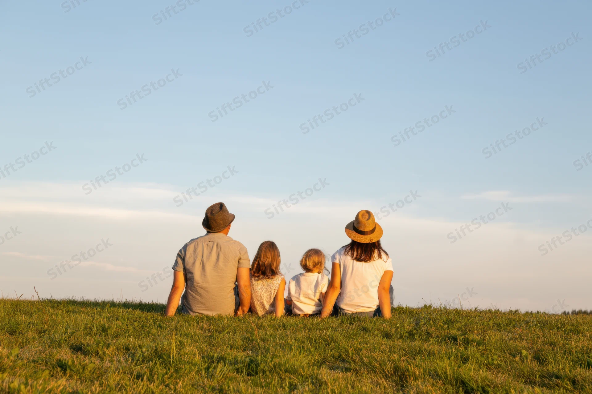 Family of four sitting on a grassy hill at sunset backs to the camera