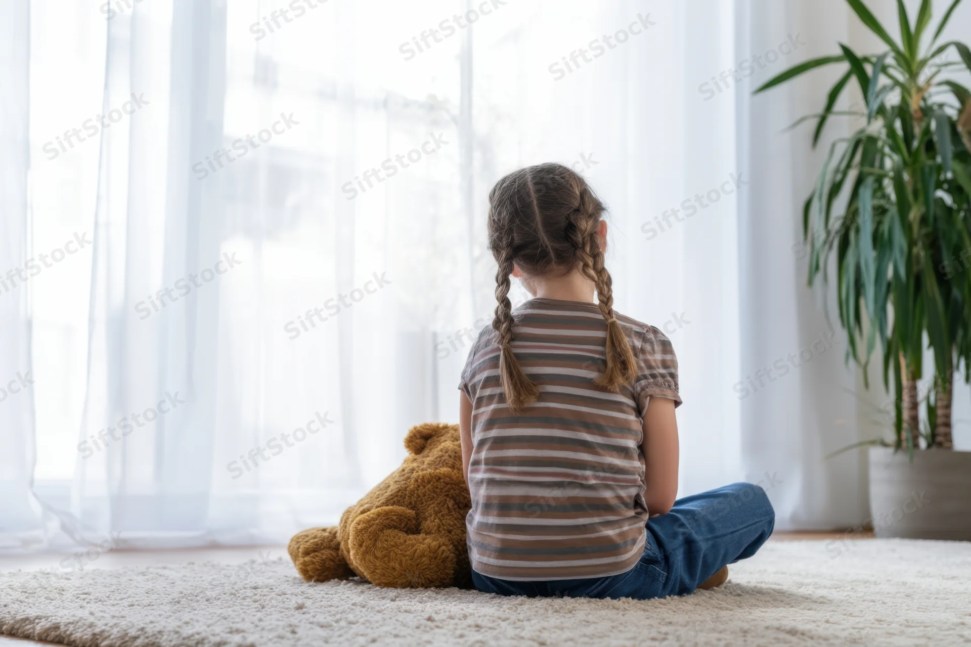 Girl with braids sitting on rug facing window with teddy bear beside her