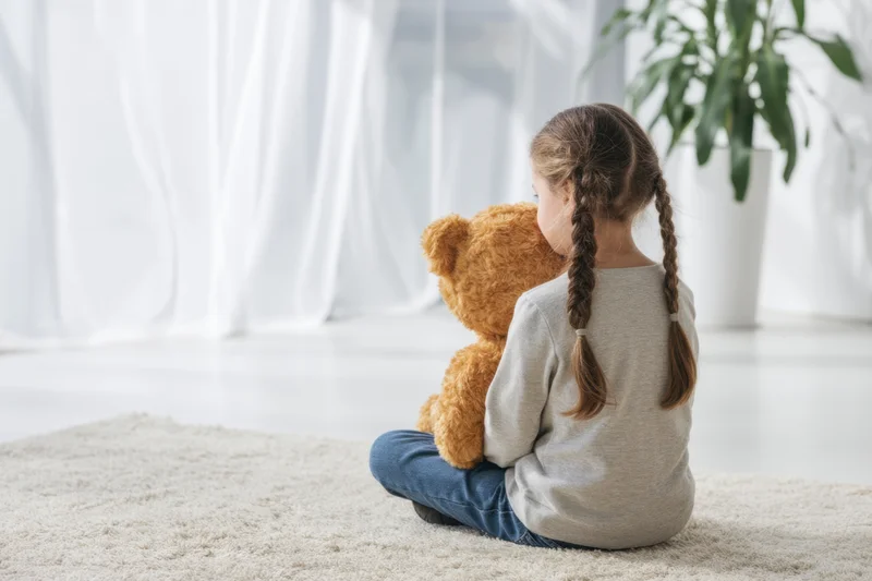 Young girl with braids sitting on a light rug holding a teddy bear, back to camera, in a bright living room