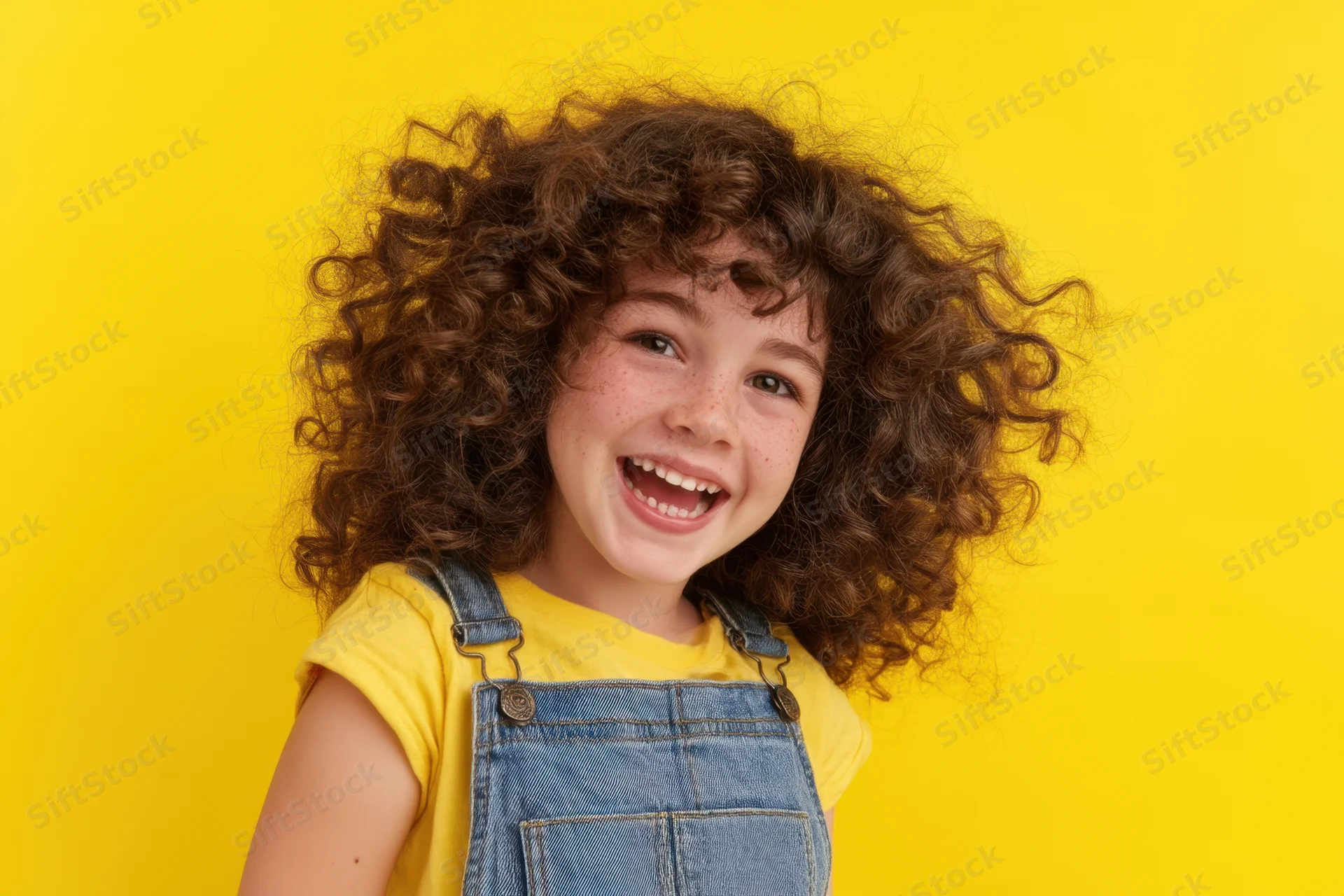 Young girl with curly hair smiling joyfully in denim overalls against yellow background