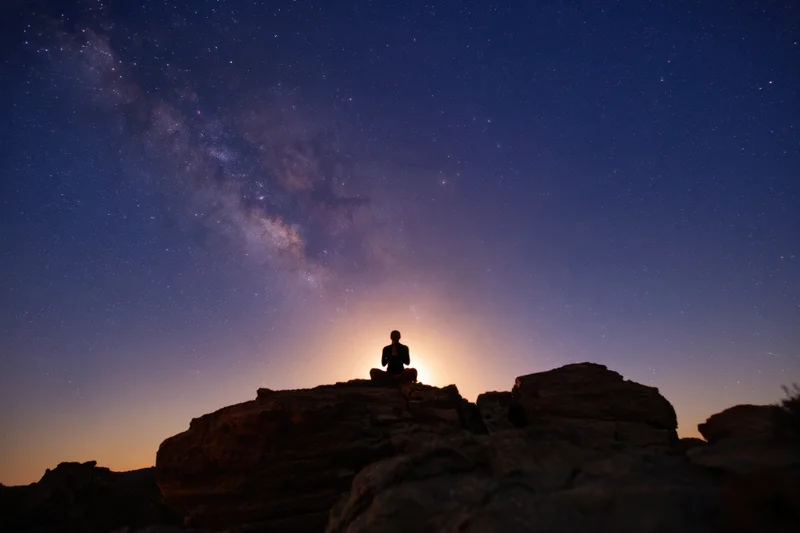 Silhouette of a person meditating cross-legged on rugged rocks under a star-filled night sky with the milky way visible above the horizon