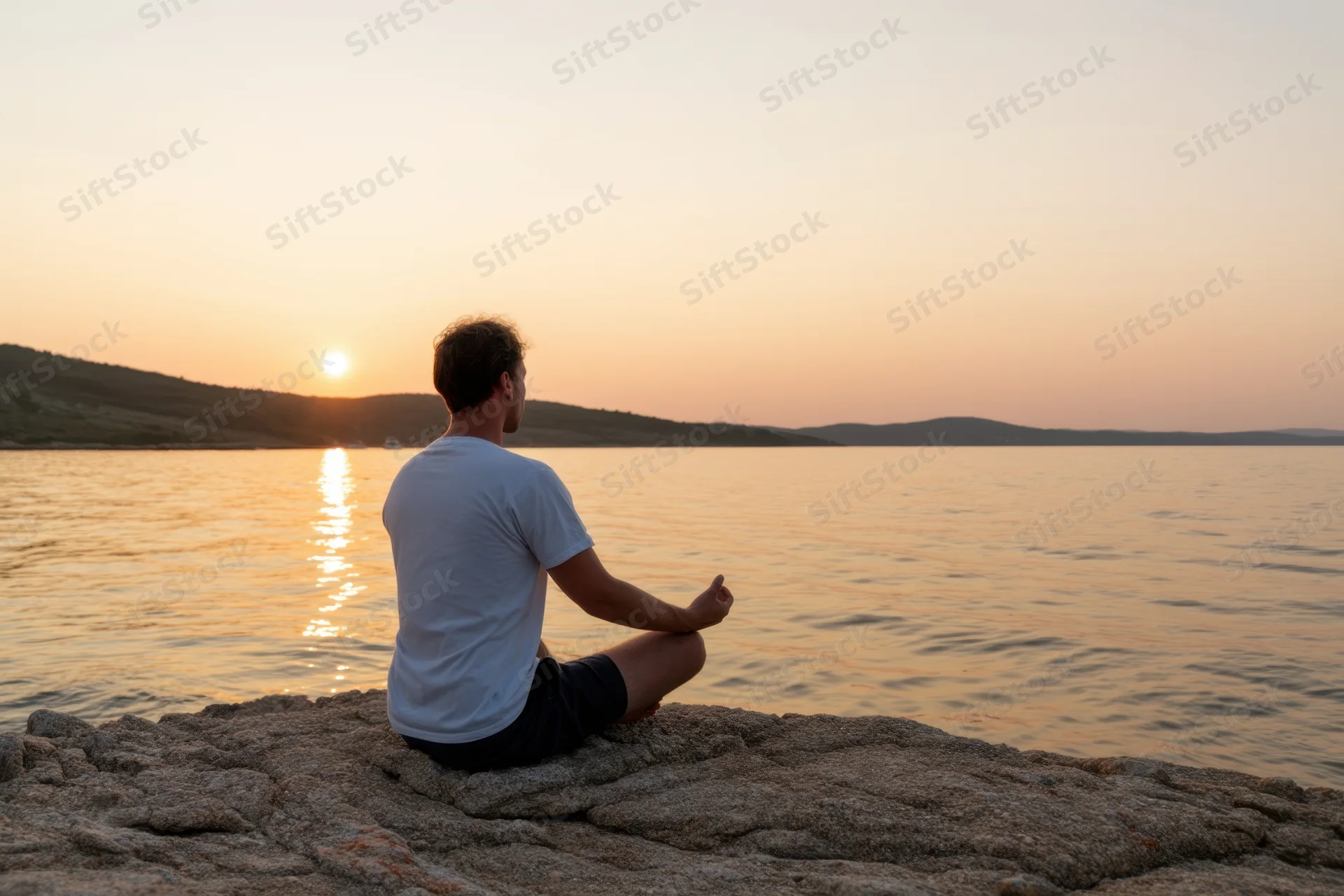 Man meditating in lotus position on a rock by a lake at sunset
