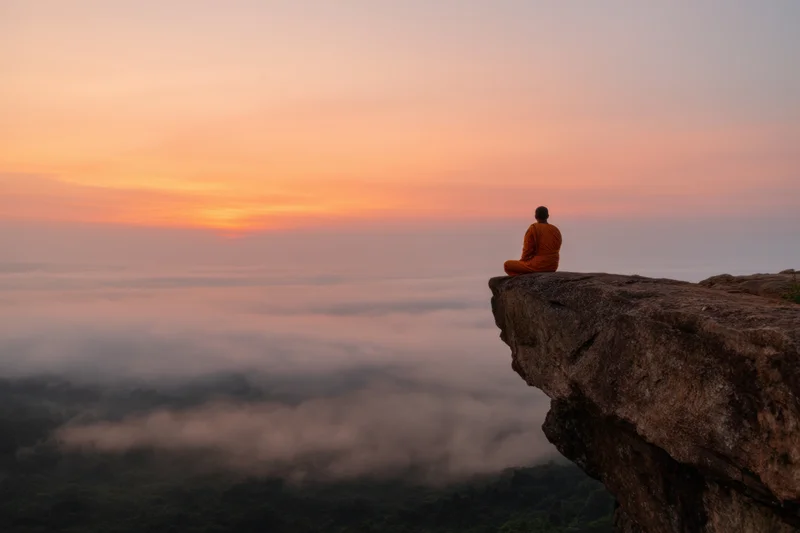 Monk meditating in saffron robes on a cliff overlooking clouds at sunset