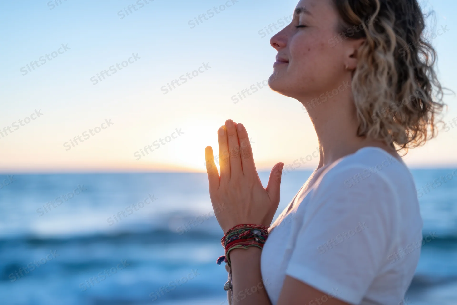 Woman in profile with hands pressed together in prayer by the ocean at sunset