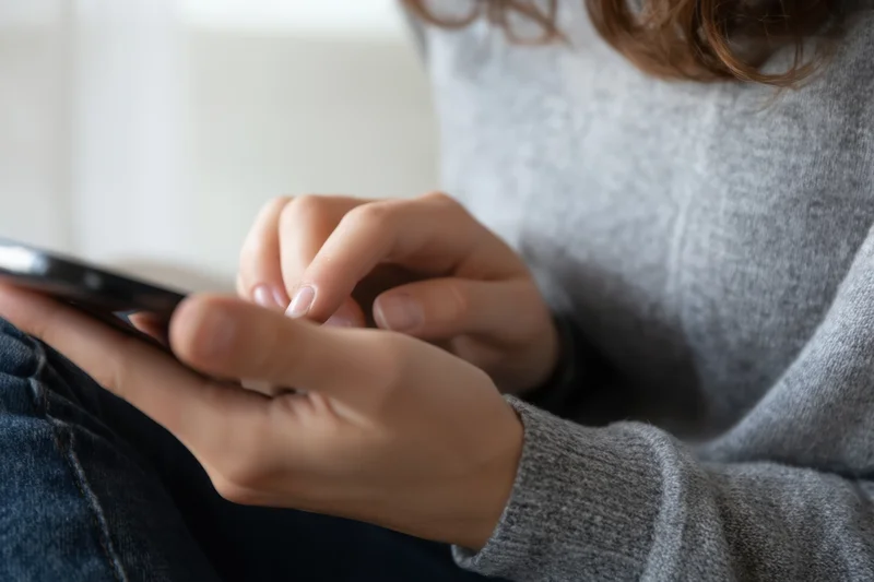 Woman tapping smartphone with hands in gray sweater and jeans indoors