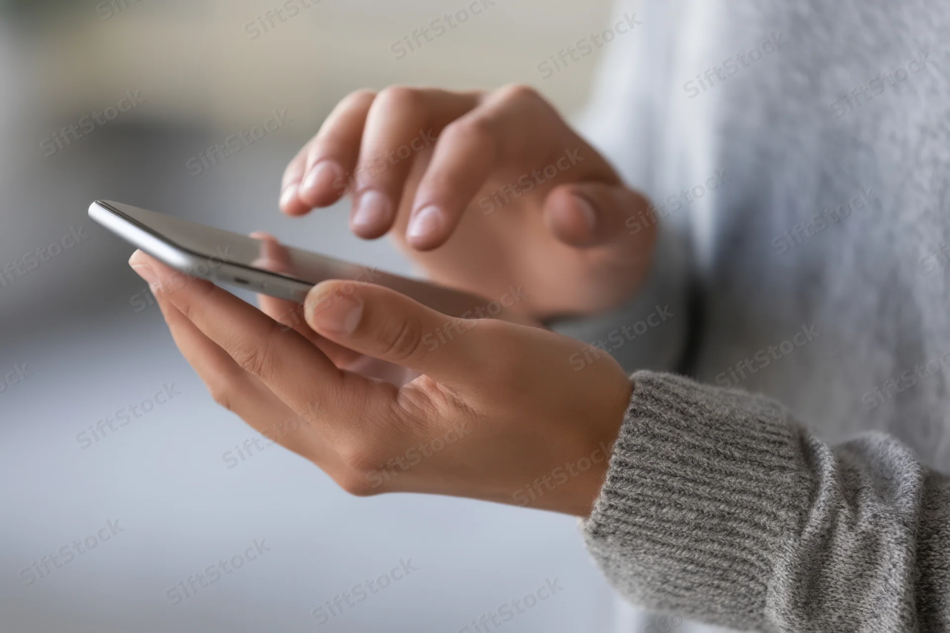Close-up of hands tapping a smartphone while wearing a gray sweater indoors