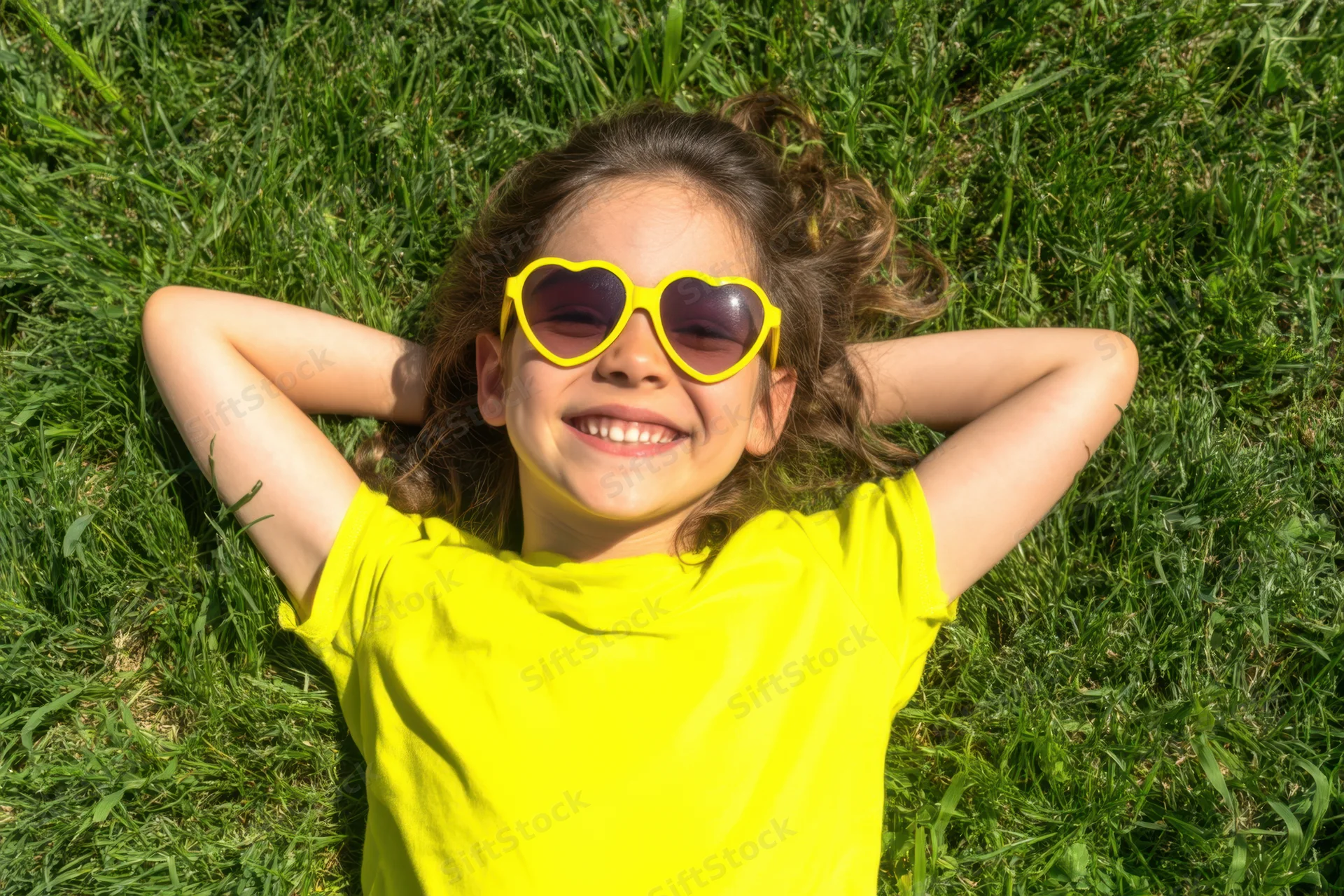 Smiling girl lying on grass with heart-shaped sunglasses and yellow t-shirt