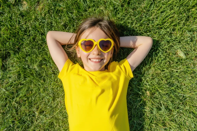 Young girl smiling while lying on green grass with hands behind head wearing yellow heart-shaped sunglasses and yellow tshirt