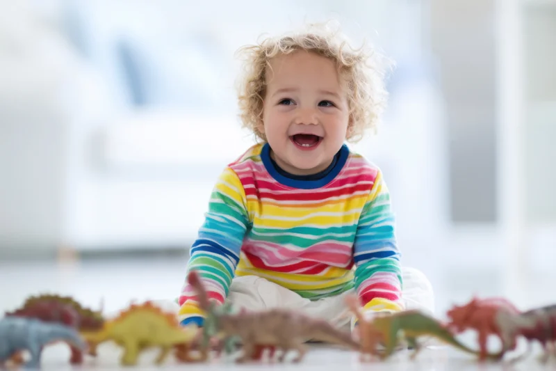 Cheerful toddler with curly blonde hair sitting on floor surrounded by colorful dinosaur toys