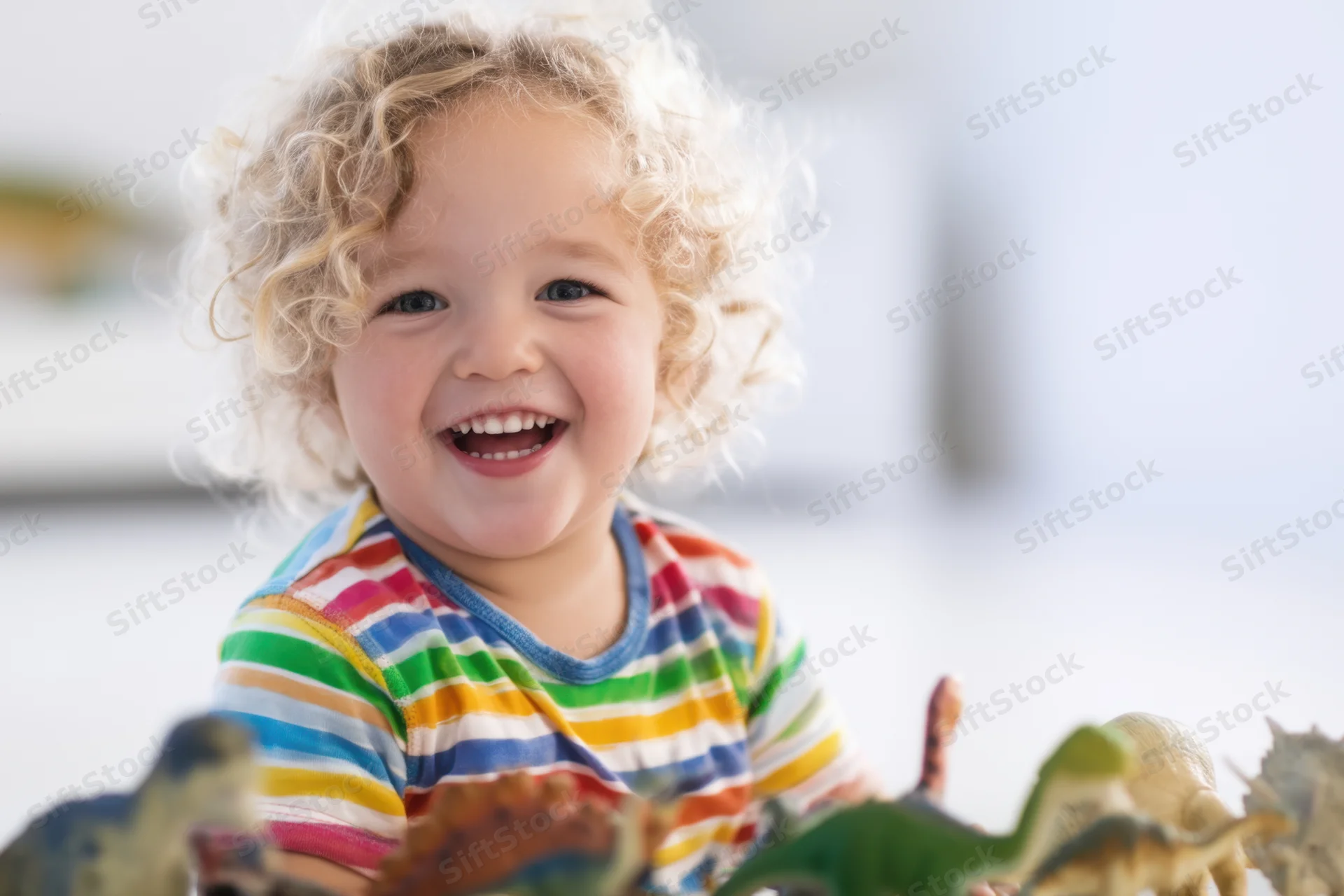 Cheerful toddler with curly blonde hair smiling while playing with dinosaur toys indoors