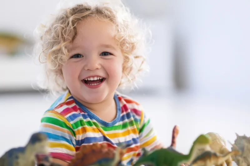 Cheerful toddler with curly blonde hair smiling while playing with dinosaur toys indoors