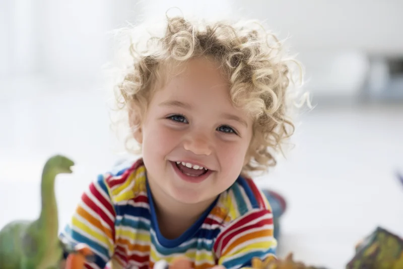 Smiling toddler with curly blonde hair playing with dinosaur toys indoors