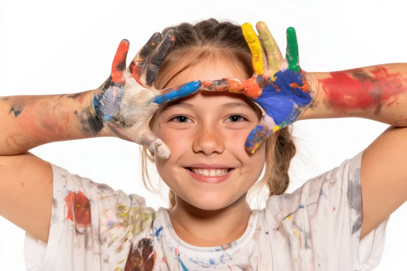 Young girl smiling with paint-covered hands and arms raised in front of her face, wearing a paint-splattered tshirt against a white background