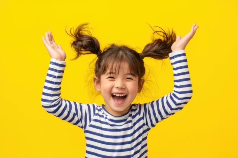 Young girl with pigtails laughing joyfully with arms raised against yellow background