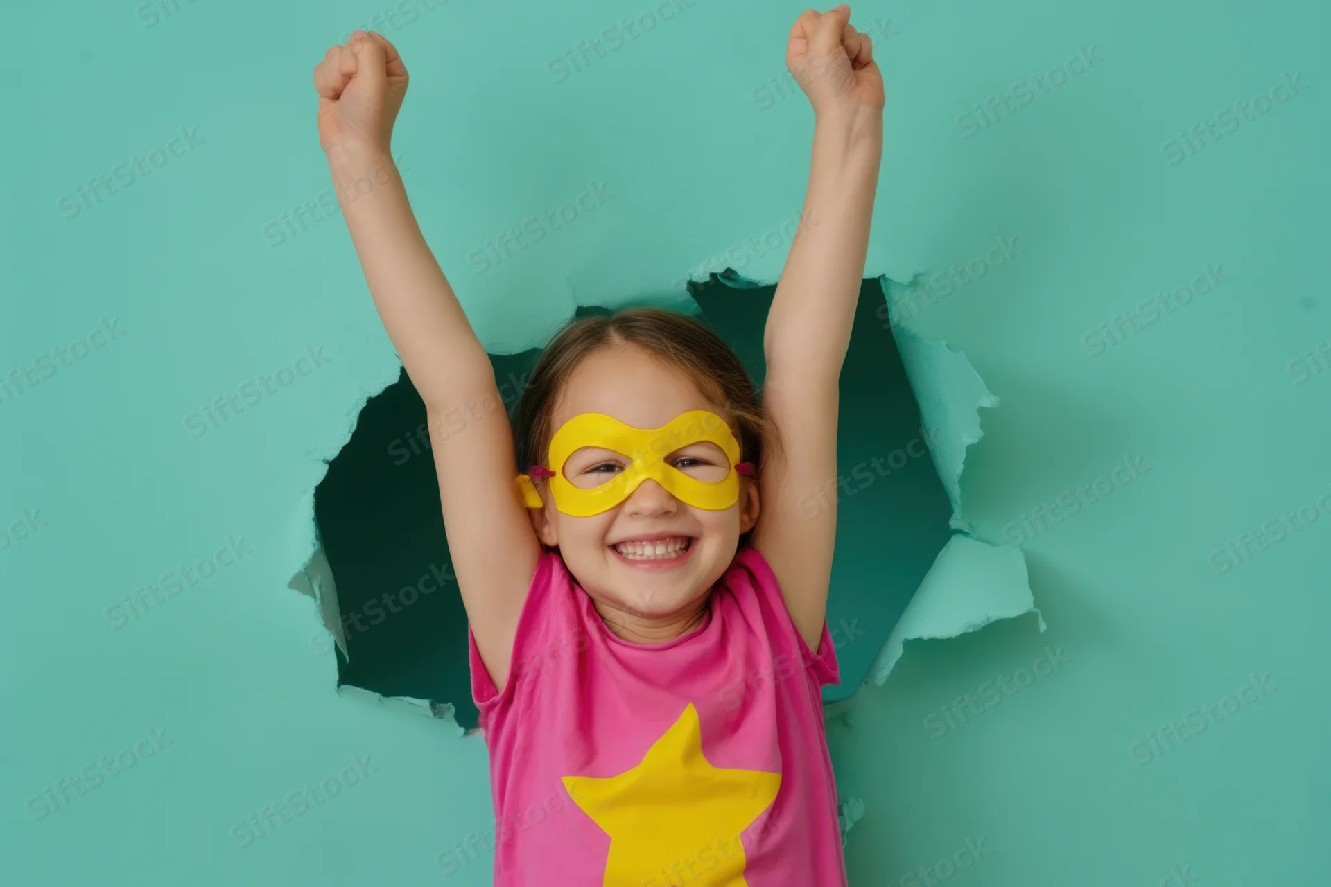 Young girl in superhero costume with yellow mask and pink shirt bursting through torn teal paper backdrop, arms raised in triumph