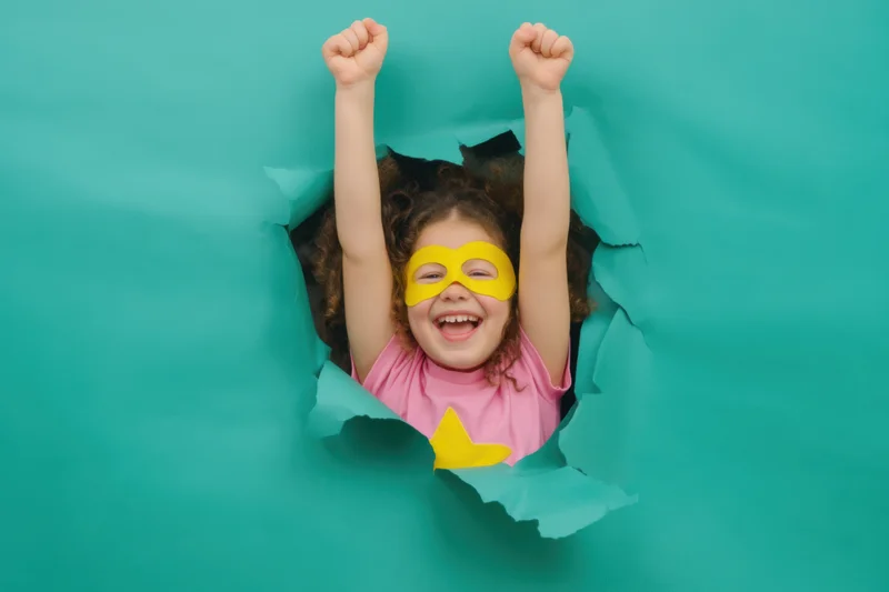 Young girl in superhero costume bursting through torn teal paper backdrop with arms raised in joy