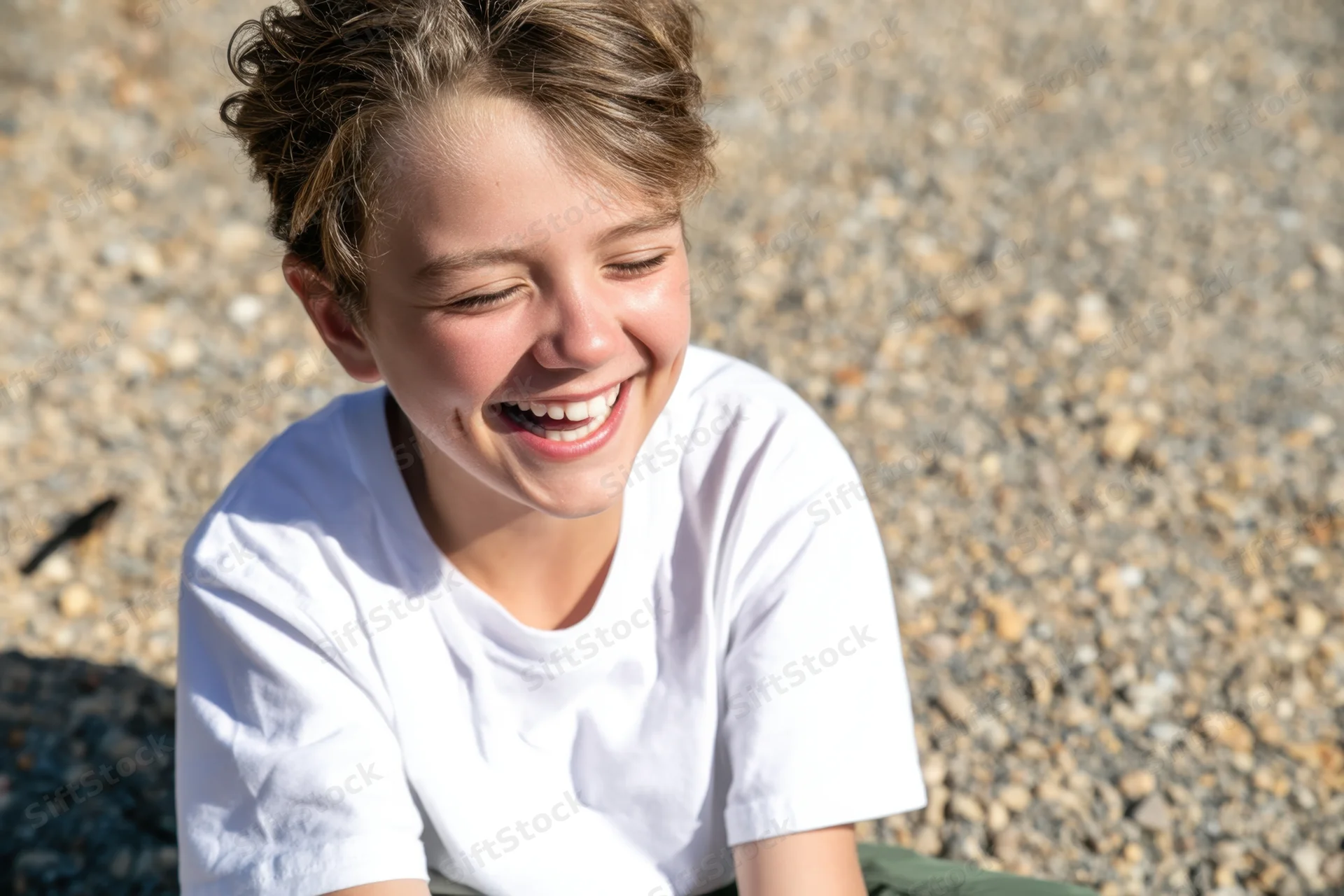 Young boy laughing with eyes closed while sitting on a gravel surface in sunlight