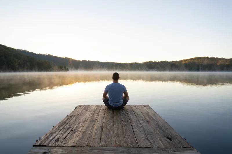 Man sitting cross-legged on wooden dock facing calm lake at sunrise