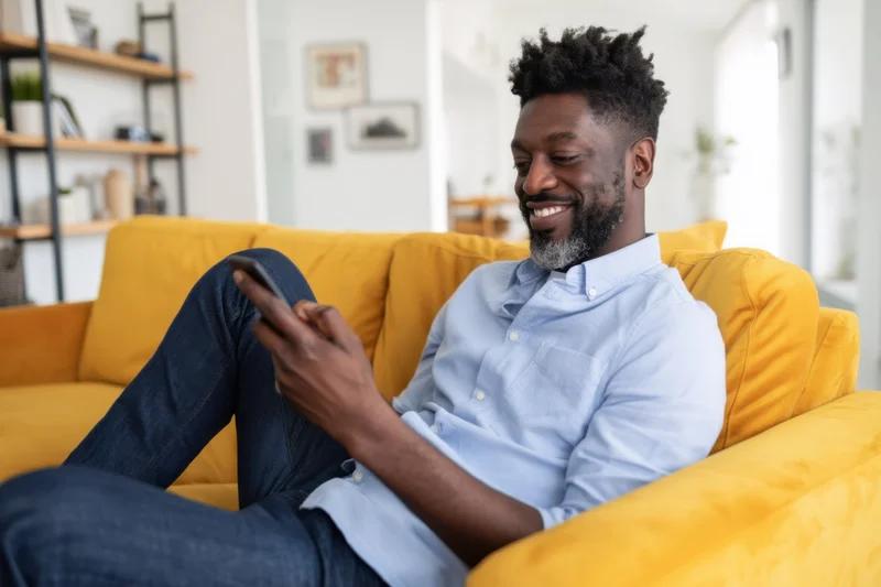 Man smiling while using smartphone reclined on mustard-yellow couch in bright modern living room