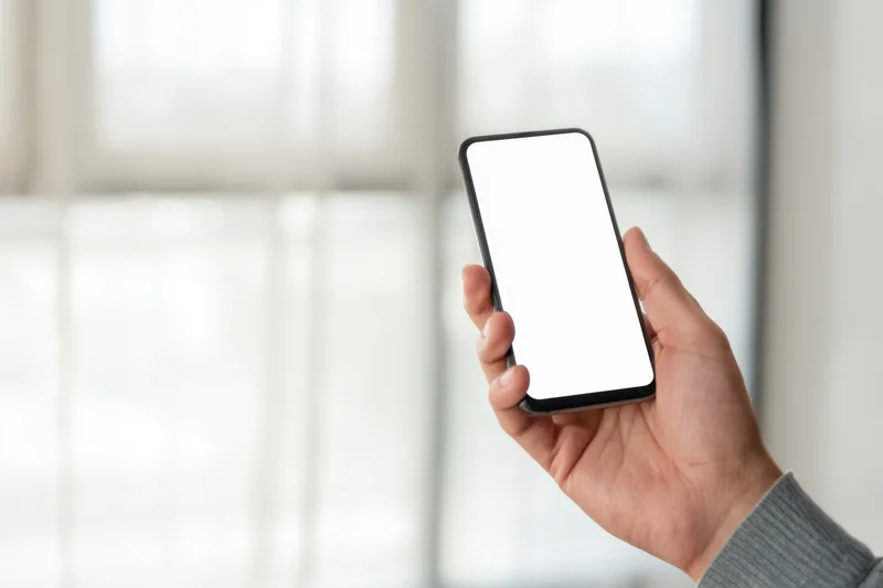 Hand holding smartphone with blank white screen indoors near window
