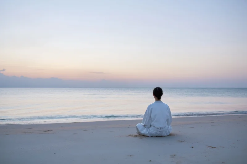 Woman sitting cross-legged on a quiet pale beach at sunset facing the ocean