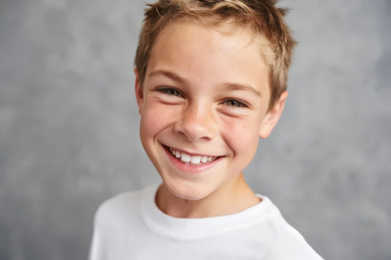 Smiling boy with short light brown hair wearing a white tshirt in front of a gray background