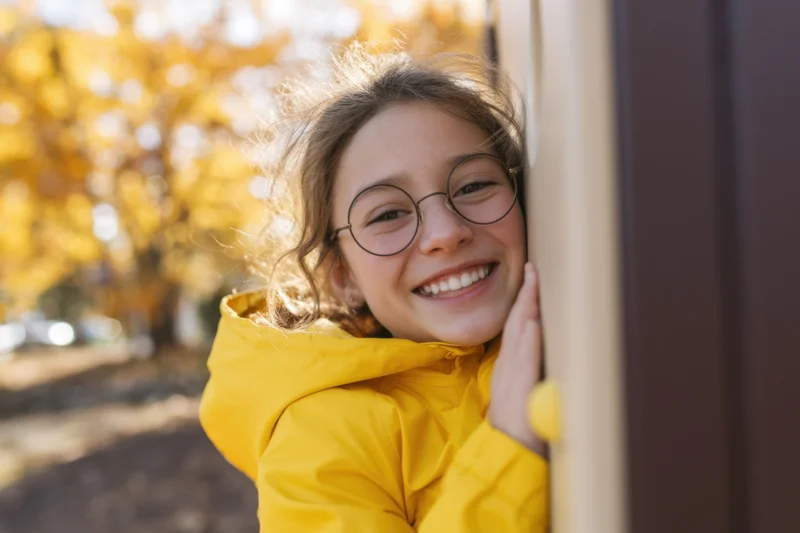 Smiling girl in yellow jacket peeking from door frame outdoors in autumn