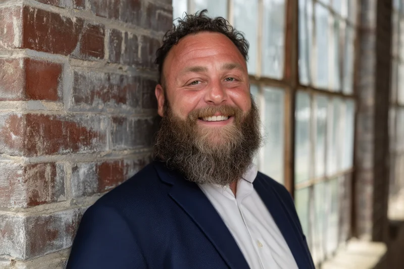 Smiling man with full beard in navy blazer leaning against brick wall near window