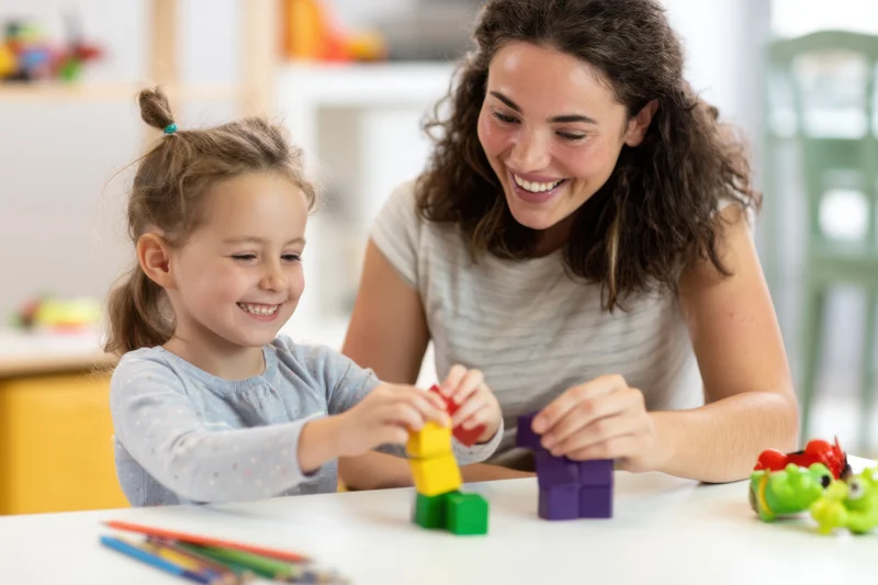 Woman and young girl smiling while stacking colorful blocks at a table in a home setting