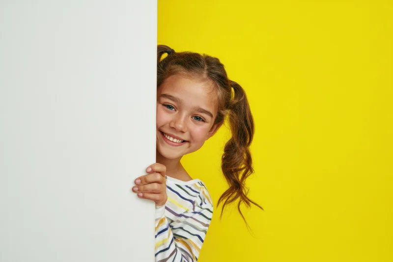 Young girl smiling and peeking from behind a white panel against a yellow background