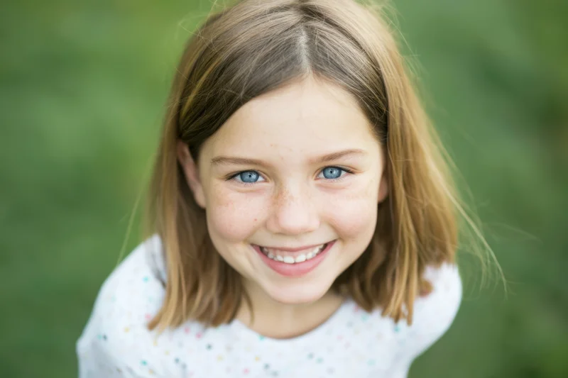 Smiling young girl with shoulder-length light brown hair outdoors in a green setting