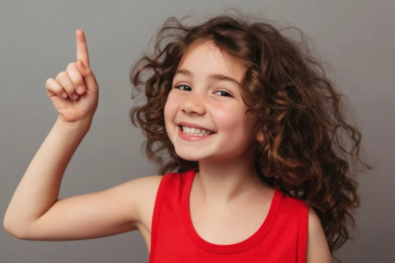 Young girl with curly brown hair smiling and pointing upward in a studio portrait