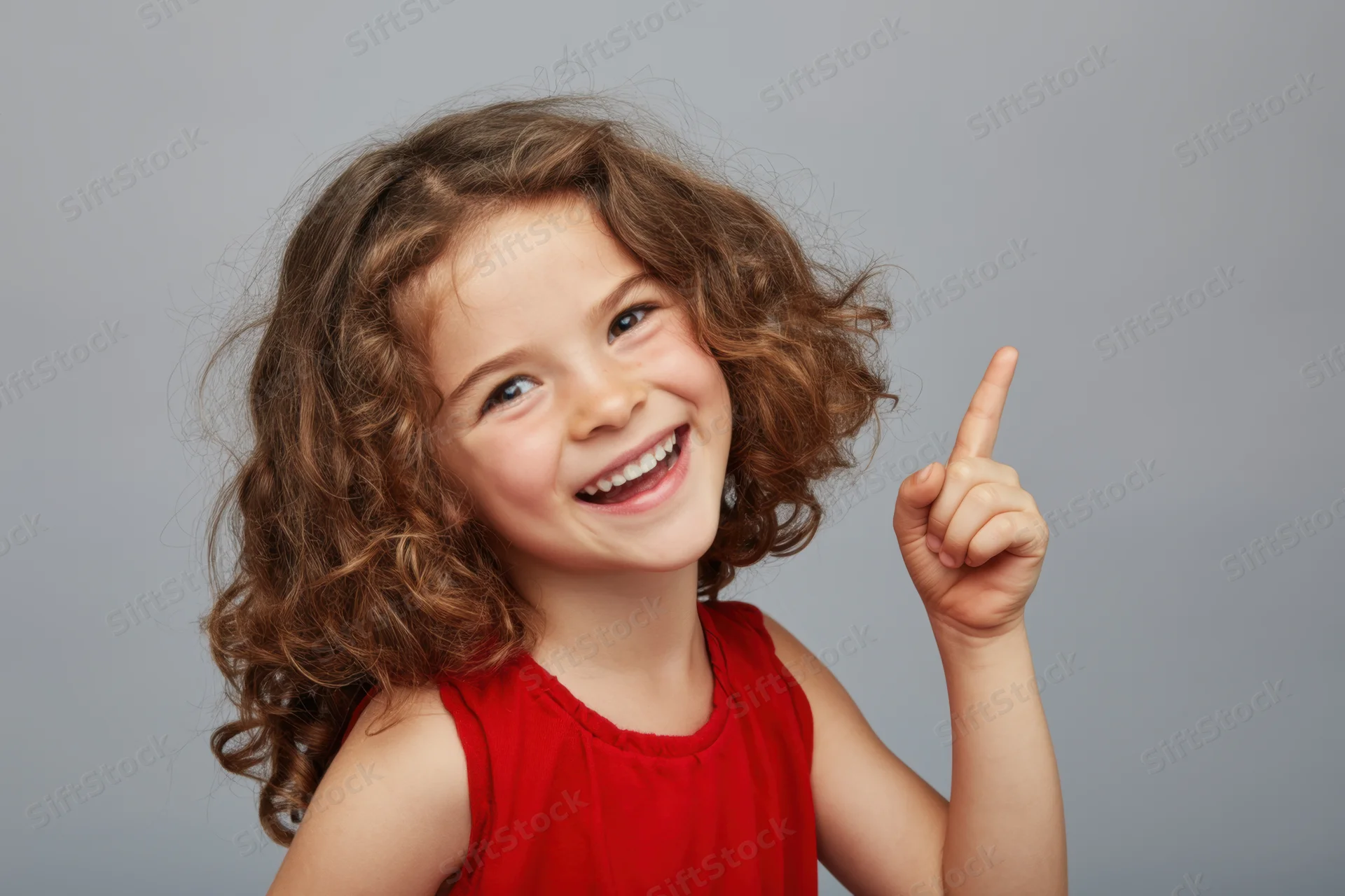 Young girl with curly brown hair smiling and pointing upward in a studio portrait wearing a red sleeveless dress