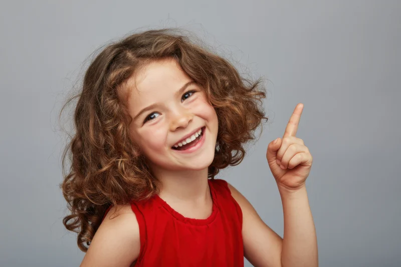 Young girl with curly brown hair smiling and pointing upward in a studio portrait wearing a red sleeveless dress