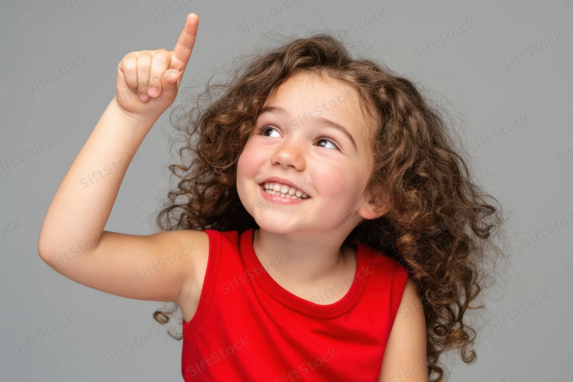 Young girl with curly brown hair pointing upward while smiling in a studio portrait
