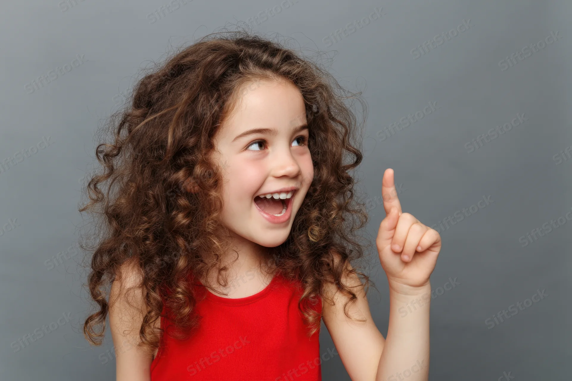 Young girl with curly brown hair smiling and pointing upward in a studio portrait