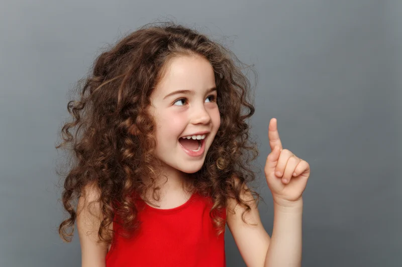 Young girl with curly brown hair smiling and pointing upward in a studio portrait