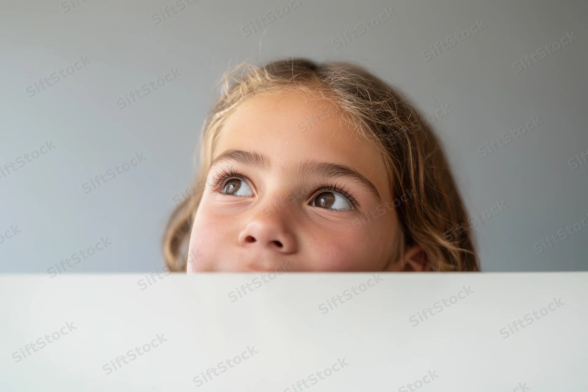 Young girl peeking over white surface with curious expression indoors